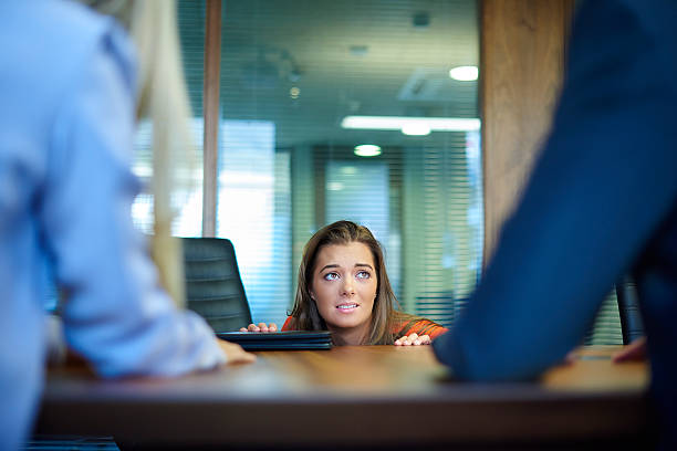 A young graduate tries to hide under the desk as she is quizzed by a job interview panel.She crouches to the edge of the desk and peeps up at the questioner.In the foreground the arms of a male and female interview board can be seen.The young woman has a cv on the desk in front of her .