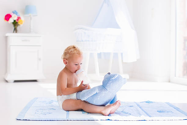 Cute blond curly baby boy wearing a diaper drinking milk on a sunny morning playing with his toy teddy bear in sunny white bedroom. Kids drink formula. Toddler with plastic bottle. Child nutrition.
