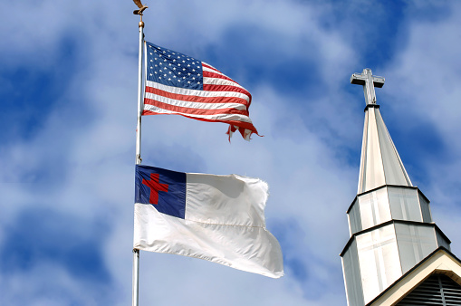 American flag and Christian flag fly besides a church steeple.  Cross seems to stand between or besides the two.