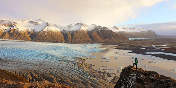 Male hiker is standing at the edge of a cliff overlooking the Skaftafell Glacier in Skaftafell National Park in Southeast Iceland. The traveller looks very small in this majestic panoramic landscape standing on top of a mountain admiring the beautiful view over one of the many tongues of Vatnajokull glacier and the adjacent lagoon.