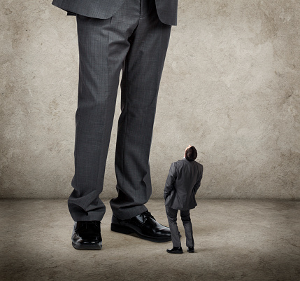 A businessman is leaning back with his hands in his pockets. He is looking up at a much larger businessman above him.  There is room for text.