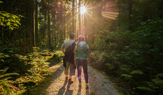 Canada:  Mature father and teenaged daughter backpacker hiking on a trail in a wilderness park stop to look at the sun's rays streaming through the trees on a summer evening.  Mount Seymour Provincial Park, North Vancouver, British Columbia, Canada.
