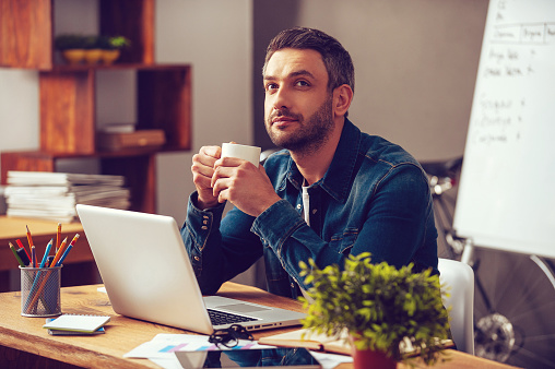 Thoughtful young man holding coffee cup and looking away while sitting at his working place in office