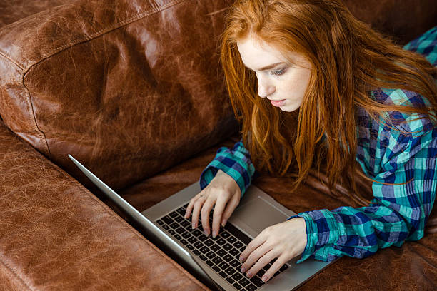 istock Concentrated redhead woman working with laptop and lying on  couch 499370844