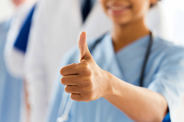 close up of doctor or nurse showing thumbs gesture, people, health care and medicine concept - close up of happy african female doctor or nurse showing thumbs up hand sign over group of medics meeting at hospital sighing stock pictures, royalty-free photos & images