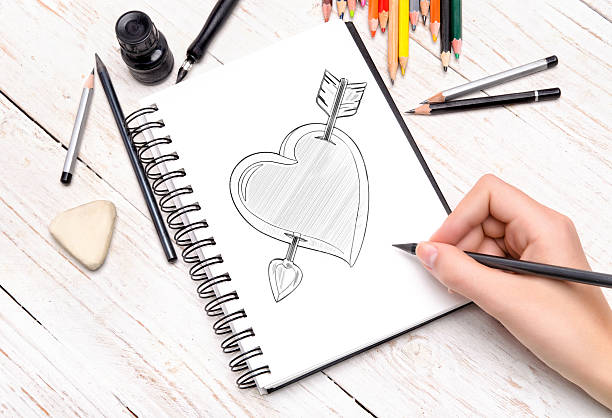 Woman's hand with decorated fingernails using a charcoal pencil to draw in a notebook on a wooden desk.