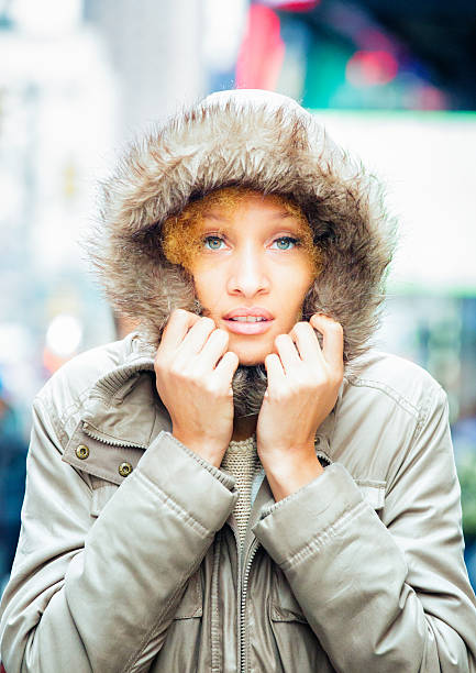 Ethereal mixed-race woman in Winter coat with a fake fur-lined cap, hit with a sudden chill. She looks ahead while holding the coat close to her face.