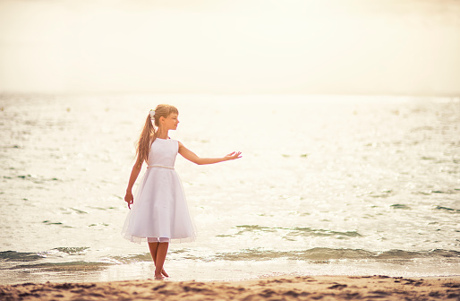 Little girl dancing ballet on the beach on sunny summer day. The girl aged 9 is wearing white dress.