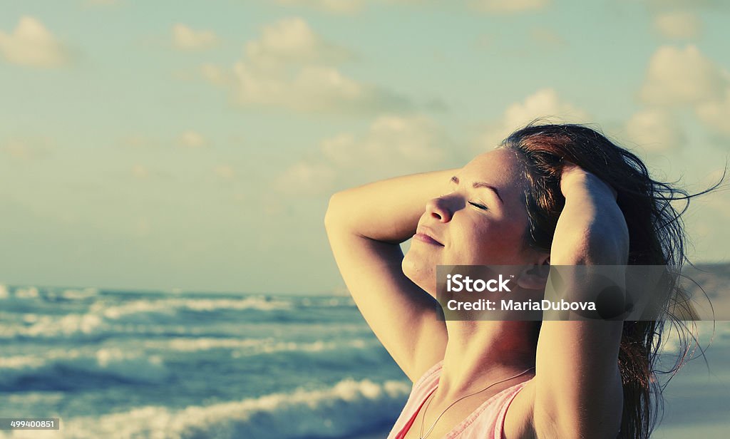 Woman at seaside with hands in hair Young woman basking in the sun on a seashore.  Only the upper part of her chest, including the arms, neck and head, is visible, while the rest of her body is obscured by the lower edge of the image.  She is wearing a pink sleeveless shirt with a thin necklace around her neck.  Her eyes are closed, and she is running her hands through her brown hair while her head is slightly tilted back.  The out-of-focus background features a sea with bubbly waves and a clear sky sprinkled with several puffy clouds.  To the right of her head, a small part of the coast can be seen. Adult Stock Photo