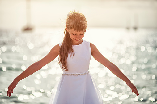 Little girl dancing ballet on the beach on sunny summer day. The girl aged 9 is wearing white dress.