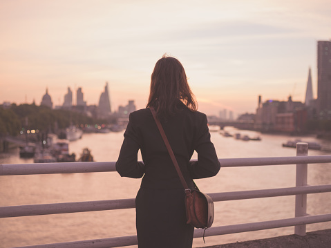 A young woman with a shoulderbag is standing on a bridge and is admiring the sunrise over the London skyline