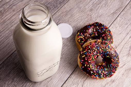 Large glass bottle of milk with two sprinkled doughnuts on the side sitting on wood planks.