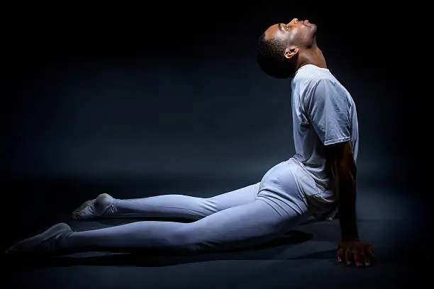 Black male dancer practicing warm up exercises for flexibility.  He is practicing on a dark studio floor.  The man is slim but tone african american.  The athletic dancer is doing yoga like poses to demonstrate the human body flexibility.  The subject is lit for extreme contrast between shadow and highlight to outline the human figure.