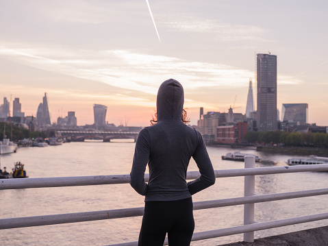 A young woman wearing a sporty hoodie is standing on a bridge in London at sunrise