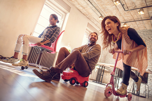 Low angle view of group of cheerful business friends having fun while behaving childish in the office. They are competing on a chair, toy car and push scooter.