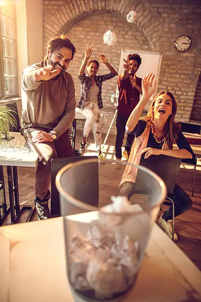 Group of happy friends having fun at casual office while throwing papers into a garbage can.
