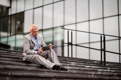 Mature businessman sitting on stairs in front of his office building and working on touchpad.
