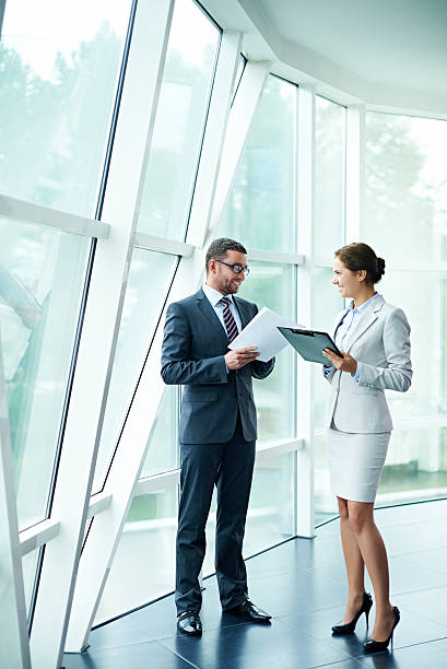 Attractive business people Two attractive business people discussing documents standing near huge window talking two people business talk business stock pictures, royalty-free photos & images