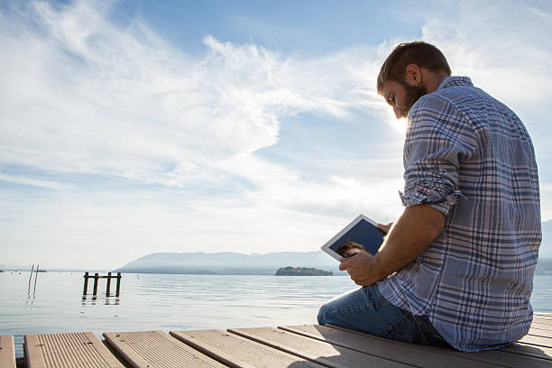 istock Young man sits on jetty above lake, uses digital tablet 499480902