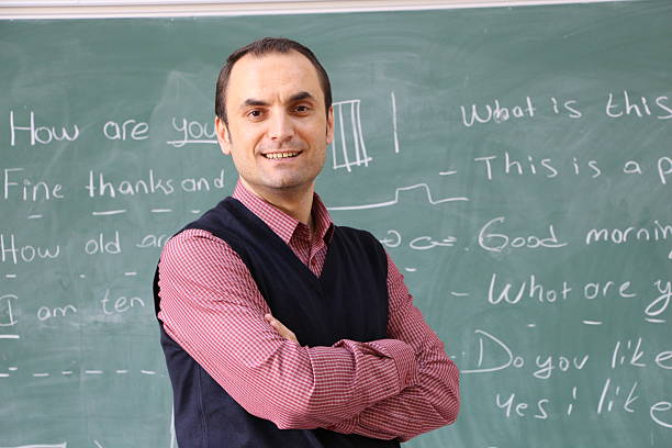 A teacher with a long white beard, big gray hair, and glasses is wearing a checkered shirt and sports jacket in front of a blackboard with complex formulas written on it. He has outstretched hands curled into fists and a happy smile on his face.
