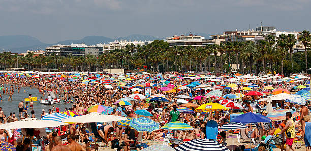 A crowded beach with colorful umbrellas, sunbathers, and palm trees, with a cityscape and mountains in the background.