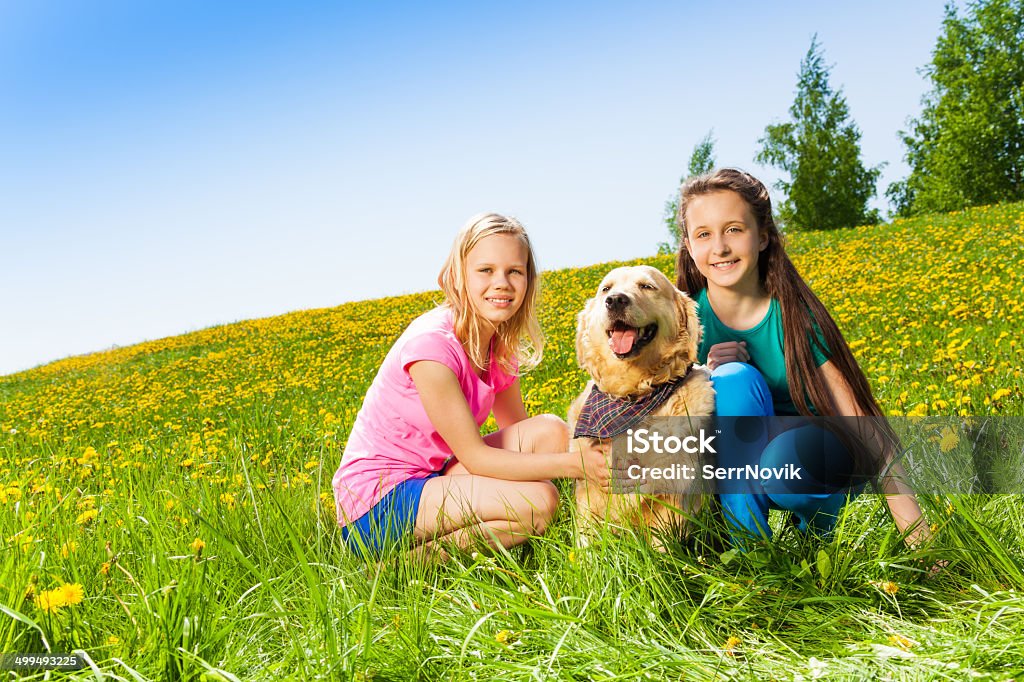 Two girls sitting near to dog on green grass Two girls sitting near to dog on green grass with yellow flowers in summer Agricultural Field Stock Photo