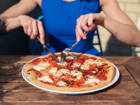 A young woman in a blue dress is sitting outside at a restaurant table and is cutting a pizza