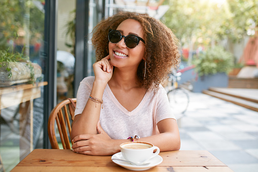 Portrait of cute young girl sitting at outdoor cafe looking at camera and smiling. African young woman at sidewalk cafe with a cup of coffee.