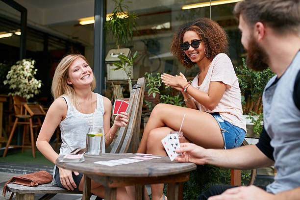 Young people at cafe enjoying playing poker game Three friends sitting in outdoor cafe and playing cards and having fun. Happy young people at sidewalk coffee shop enjoying playing poker game. friends playing cards stock pictures, royalty-free photos & images