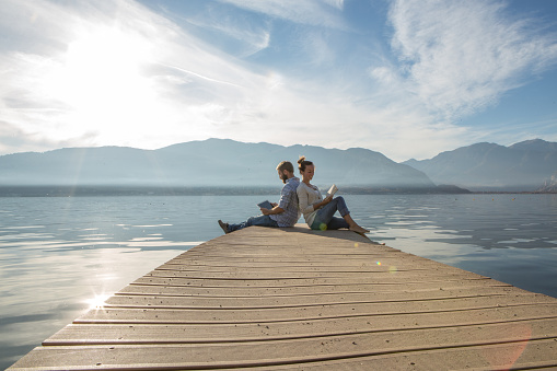 Young couple sits on a jetty above the lake, uses a book and a digital tablet. Beautiful Autumn day in Italy.