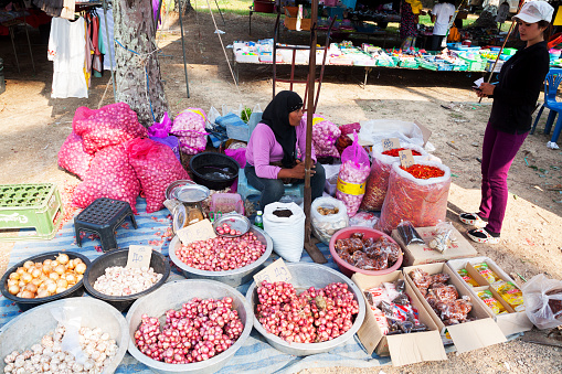 Ao Nang, Thailand - March 9, 2013: A thai woman with headscarf is selling onions and garlic on local market in outskirts of Ao Nang. Woman is sitting on floor under parasol. In background are fashion market stalls.