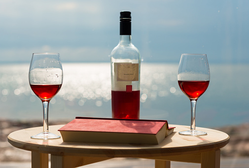 Rosé wine glasses and bottle next to red book on table next to the sea