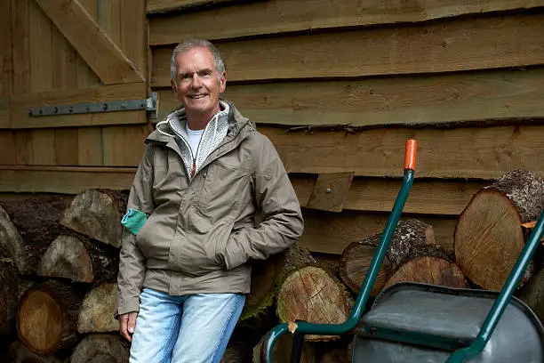 Portrait of confident senior man standing against stacked firewood outside cottage