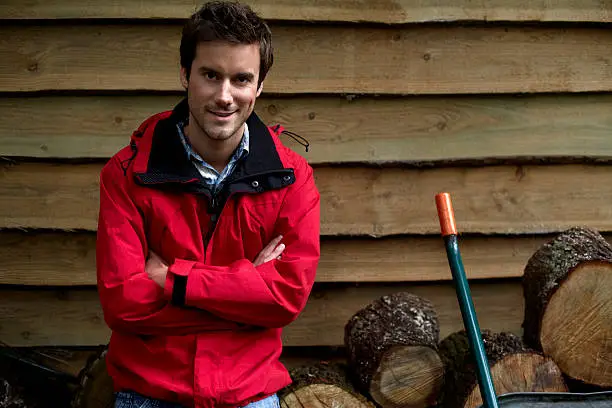 Portrait of confident young man in winter coat standing against firewood outside cottage