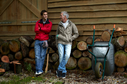 Full length of father and son conversing while standing by against firewood outside cottage
