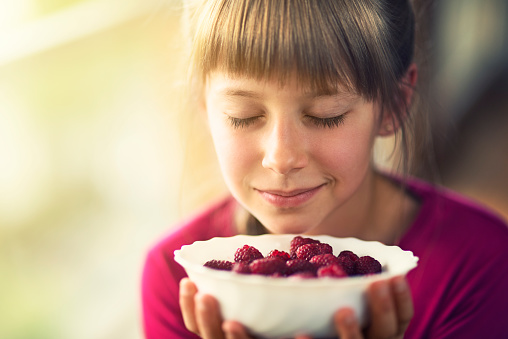 Smiling little girl eating raspberries. Closeup of the beutiful little girl smelling raspberries. She is wearing a red blouse and holding a white bowl of raspberries. She is sitting next to the window.