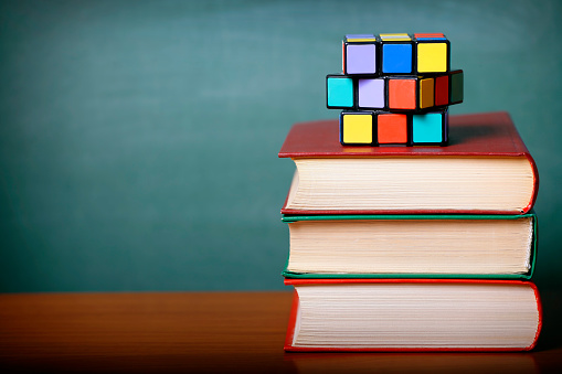 Ulyanovsk, Russia - November 30, 2015: Stack of books and a Rubik's cube on a wooden desk. Green chalkboard background. Shallow depth of field. Space for copy. Can illustrate the concept of education, logic, learning, etc.