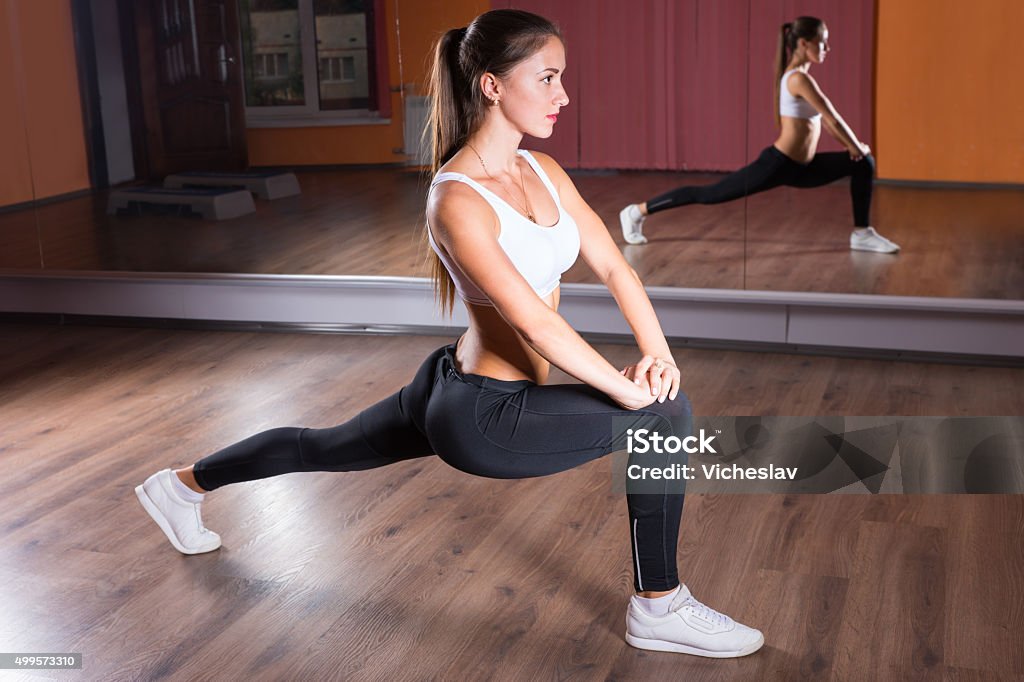 Young Woman Stretching in Lunge Position Full Length Profile View of Young Brunette Woman Wearing Exercise Clothing and Stretching in Leg Lunge Position with Straight Back in Dance Studio in front of Mirrors 2015 Stock Photo