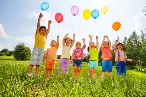 Happy kids with balloons and arms up in the sky in green field