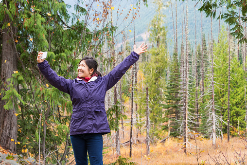 Canada:  An exuberant, lone, multi-ethnic (Asian/Caucasian) teenager takes a break from hiking to pose for a selfie on her mobile phone in a wilderness area.  Trees turning colour in the foreground and mountain in the background on an autumn day.  Near Whistler, British Columbia, Canada.