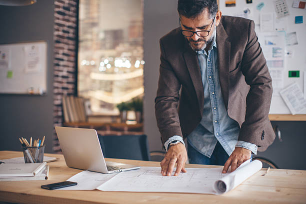 Man working at modern office space. Mature businessman working at modern office space. Standing near the table, looking at project or architect blueprint. Wearing casual clothes. Caucasian ethnicity. design occupation stock pictures, royalty-free photos & images