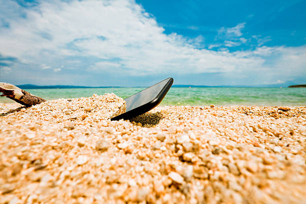 Thrown, half buried in the sand of an ocean or sea shore sand smartphone. Nobody, blue sky with some clouds above.