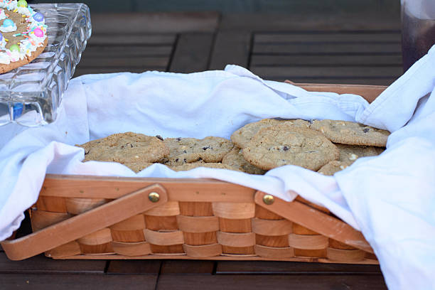 Chocolate chip cookies in a woven basket on an outdoor wooden table with a decorated cookie on a glass platform to the side.