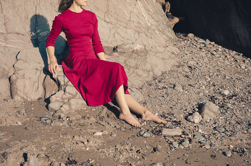 A young woman wearing a red dress is relaxing by the entrance to a cave on the beach