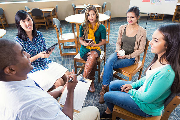 Diverse group of teen and young adult women are sitting in a circle during a meeting or support group therapy session. Young women are listening to mid adult African American male advisor or counselor. Women are holding digital tablets or coffee while listening.