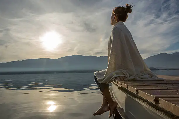 Photo of Young woman relaxes on lake pier with blanket, watches sunset