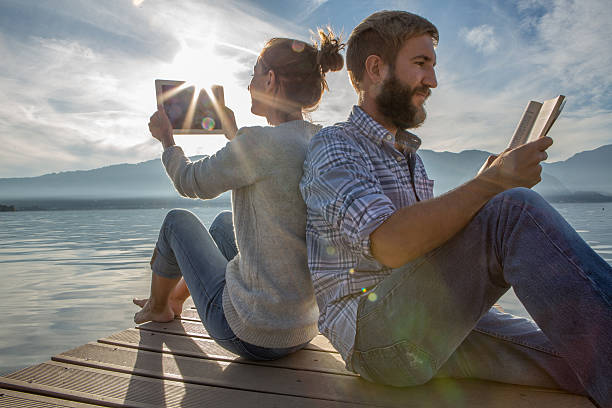 istock Young couple sits on jetty above lake, uses book/digital tablet 499659372
