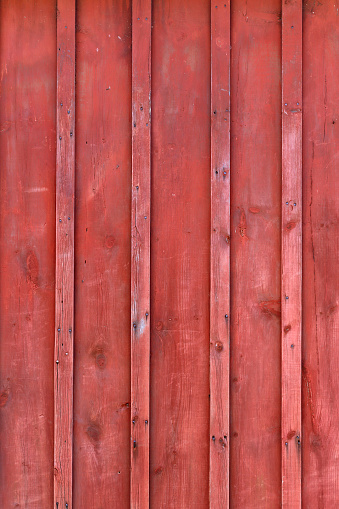 The side of an old farm building's red peeling paint rustic barn-board plank wall background with board and batten siding.