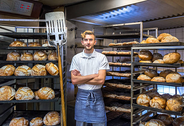 Baker in his bakery baking bread baker posing in his bakery bakehouse in the early morning between fresh baked artisan bread bakery photos stock pictures, royalty-free photos & images