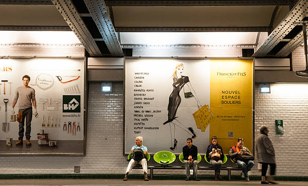 Paris, France - May 14, 2012: Four persons sit and one senior man walking along the platform of an underground station in Paris,capital of France.Huge colorful billboards behind the passenger waiting for the tube